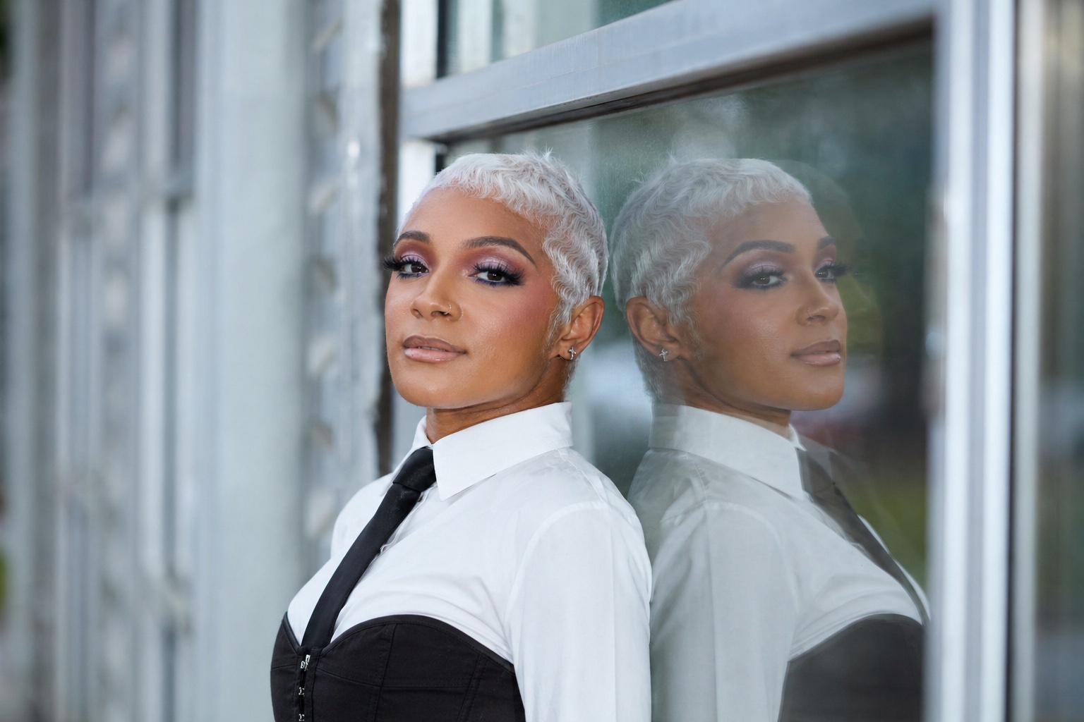 Model with a platinum pixie cut in a white shirt and black tie, reflected in a glass storefront